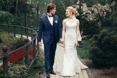 şık gelin ve damat Park'ta rahatlatıcı. yürüyüş ve ağaçlar arasında gülümseyen mutlu lüks düğün çifti. adam mavi takım elbise ve resepsiyon açık havada, beyaz elbiseli kadın