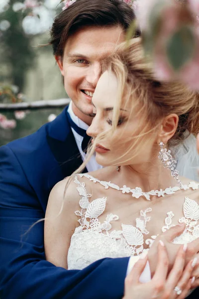 スタイリッシュな花嫁と花婿のモクレンの花の公園を受け入れます 情熱的な豪華な結婚式のカップル笑顔とハグします ロマンチックな官能的な瞬間 — ストック写真