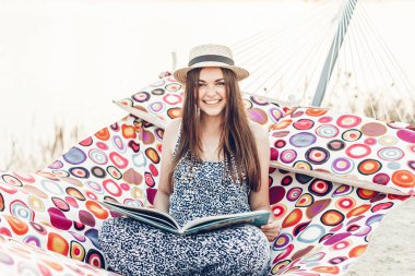 Bir hamakta, gün batımında sahilde istirahat ederken şık şapka okuma kitabı duygusal kız rahatlatıcı güzel hipster kadın