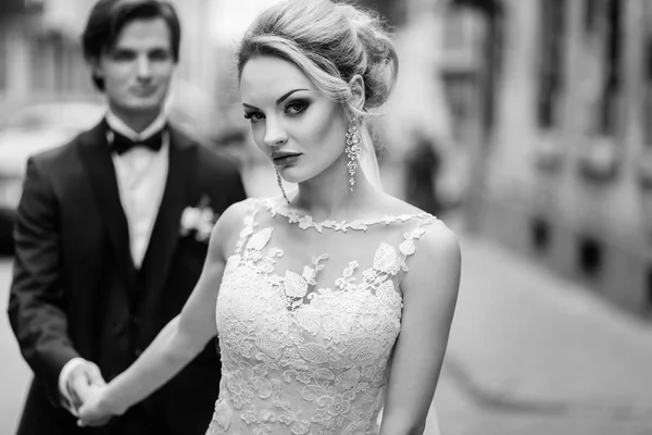 スタイリッシュな花嫁と花婿の街の通りを歩いてします 豪華な結婚式のカップル古い建物の光の中で手を繋いでいます ロマンチックな官能的な瞬間 女性を見ていると人間のポーズ 黒と白 — ストック写真