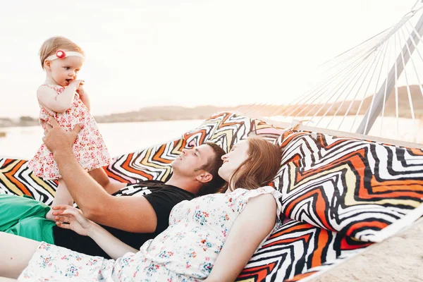 Strong young father holding his baby daughter in hands with happy hipster mother, smiling family relaxing in a hammock on a beach near lake at sunset, family vacation concept