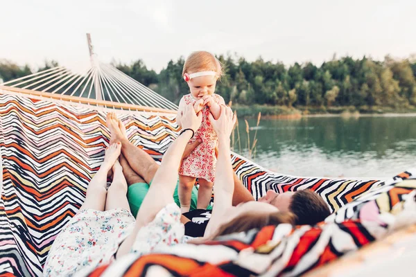 Cute baby girl in stylish dress with her parents lying in a hammock near lake and forest on family vacation trip, adorable hipster family with daughter