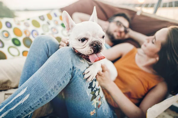 happy hipster couple with bulldog relaxing in hammock on the beach in sunset light, summer vacation. stylish family with dog cuddling and resting, cute moments. space for text
