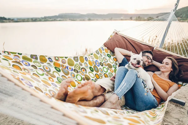 Hipster family on vacation concept, happy woman and man relaxing on a hammock at the beach with their cute bulldog pet, couple lying near a lake at sunset