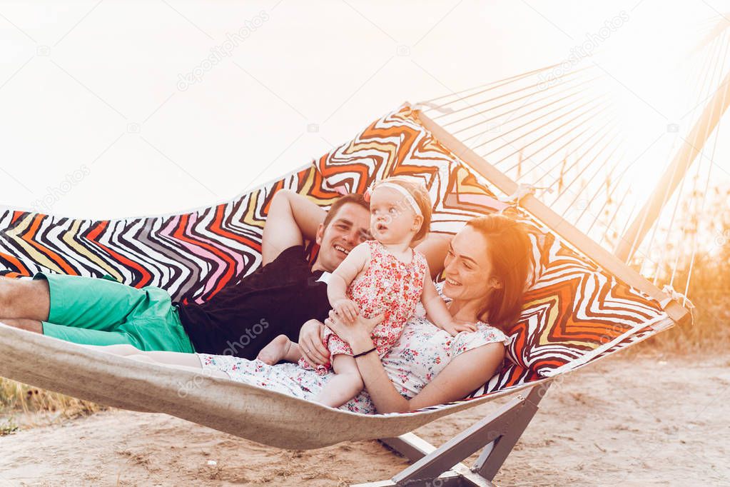 happy stylish family playing with cute daughter, relaxing in hammock on summer vacation in evening sun light on the beach. hipster couple with child resting in sunset. space for text. summer holiday