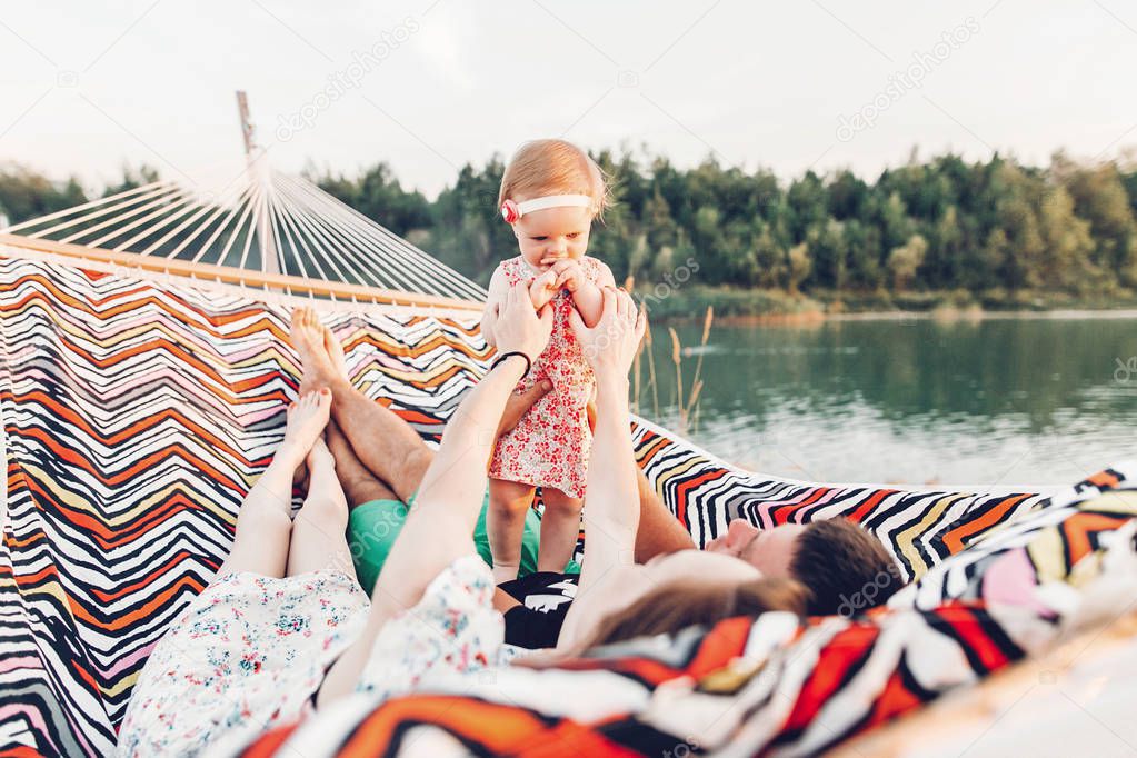 Cute baby girl in stylish dress with her parents lying in a hammock near lake and forest on family vacation trip, adorable hipster family with daughter