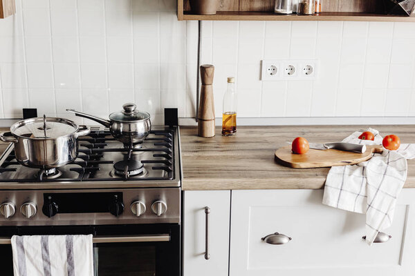 деревянная доска с ножом, помидорами, оливковым маслом на современной кухонной столешнице и стальной плите, а также полка со специями и растениями. приготовление пищи. Стильный дизайн интерьера кухни в скандинавском стиле
