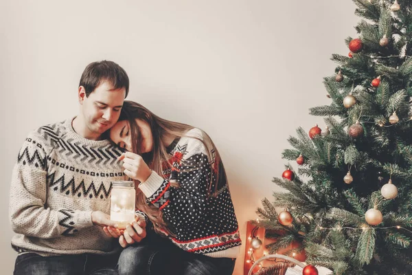 クリスマス ツリーと採用お祝いルームでランタンの光を保持しているスタイリッシュなセーターで幸せなカップル 大気中のお祝い瞬間 メリー クリスマスと新年あけましておめでとうございますコンセプト — ストック写真