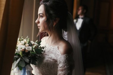 muhteşem gelin buketi ve şık damat vasıl pencere lüks otel odasında poz ile şaşırtıcı elbiseli. zengin düğün çift ışık. Yeni evliler kapalı klasik romantik anı