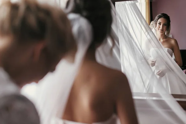 ブライドメイドと母親支援のゴージャスな花嫁の結婚式のための準備します ドレッシング ガウン リボンを押しながら花嫁が窓の近くの鏡で見ている美しい女性 — ストック写真