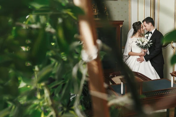 ゴージャスな花嫁ブーケとスタイリッシュな新郎がホテルで豪華な部屋で緑の葉の中で優しく抱き締めます 豊富な結婚式のカップルを受け入れます 屋内での古典的な新婚夫婦の官能的な瞬間 — ストック写真