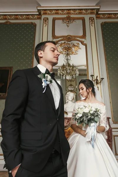 花束とスタイリッシュな新郎の高級ホテルの部屋でポーズをとって素晴らしいドレスにゴージャスな花嫁 豊富な結婚式のカップル 屋内での古典的な新婚夫婦のロマンチックな官能的な瞬間 — ストック写真