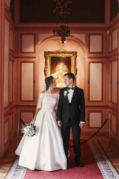 花束とスタイリッシュな新郎の高級ホテルの部屋でポーズをとって素晴らしいドレスにゴージャスな花嫁 豊富な結婚式のカップル 屋内での古典的な新婚夫婦のロマンチックな官能的な瞬間 — ストック写真