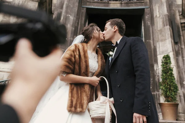カメラマン コート スタイリッシュな新郎挙式後の教会のドアの近くで豪華な花嫁の写真を撮影 幸せな新婚のカップルがキスします ロマンチックな瞬間 — ストック写真