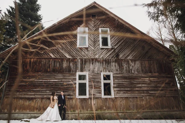 华丽的新娘在大衣和时尚的新郎摆在木房子在冬季森林 快乐的新婚夫妇轻轻拥抱在雪地公园 谷仓婚礼 浪漫的感性时刻 — 图库照片
