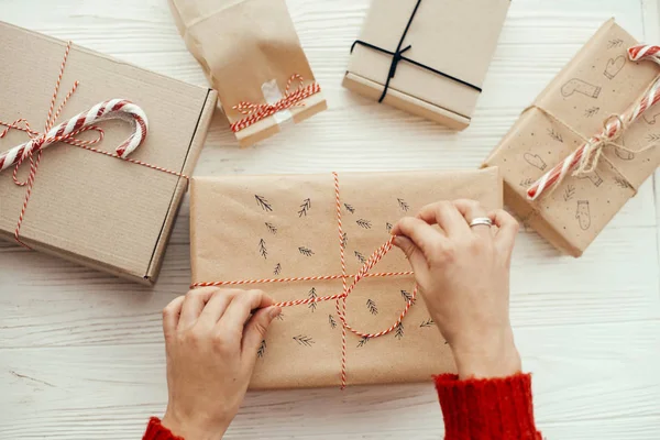 woman in red sweater wrapping stylish christmas gift with red robe. modern craft presents on white wooden background. merry christmas concept. flat lay.  seasonal greetings