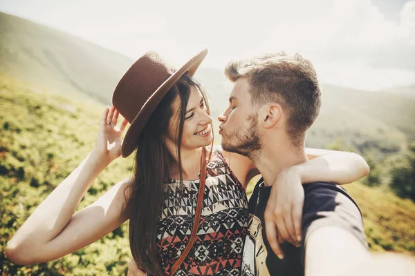 スタイリッシュな流行に敏感なカップル Selfie をとり 日当たりの良い夏の日に山の上にキスします 美しい男と新婚旅行に帽子を受け入れ幸せな女 一緒に旅行します — ストック写真