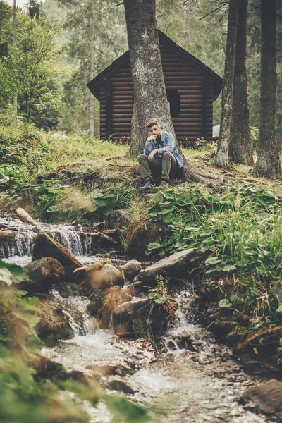 时髦的旅行者坐在小屋和河附近在晴朗的森林和山 旅行和流浪概念 文本的空间 快乐的时髦家伙在树林里放松 — 图库照片