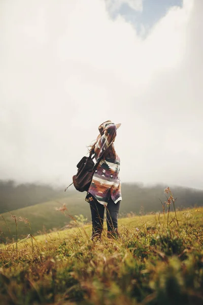旅行和流浪概念 时尚的旅行者时髦的女孩拿着帽子 带着背包和刮风的头发 在山里漫步在云端 文本的空间 惊人的大气时刻 — 图库照片