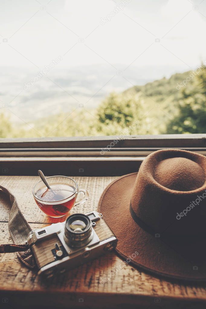 summer travel and wanderlust. stylish hipster hat, photo camera and tea in glass with spoon on wooden table at window light with view on mountains and sky. delicious hot drink on top