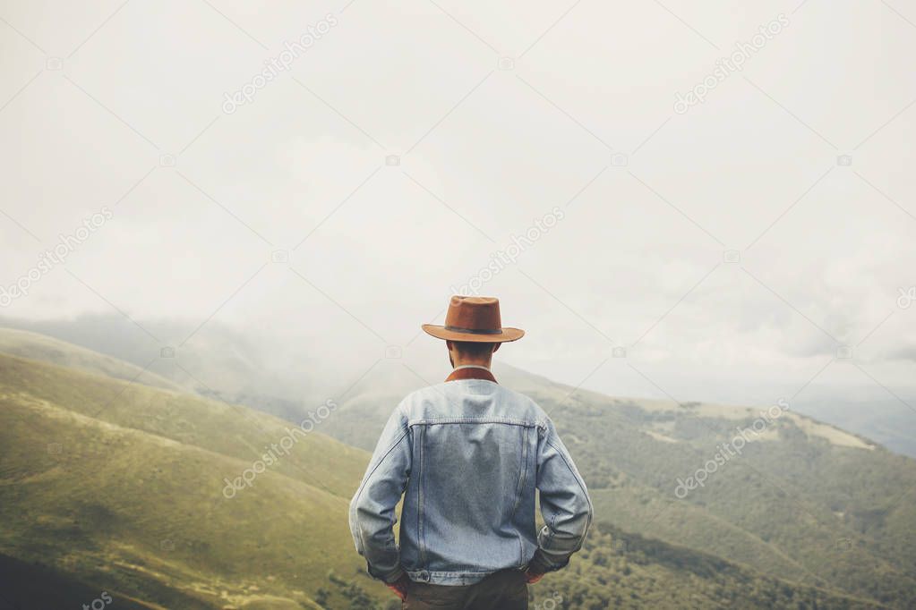 travel and wanderlust concept. stylish traveler man in hat standing on top of sunny mountains in clouds. space for text. hipster guy traveling. amazing atmospheric moment.