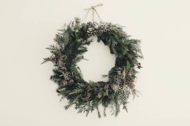 Modern Noel çelengi. Beyaz duvarında çam kozalakları, köknar dalları ve kar asılı şık bir kır çelengi. Metin için boşluk. Kış tatilleri için el yapımı dekor