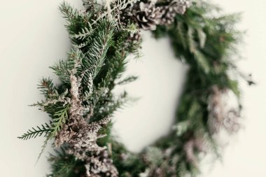 Modern Noel çelengi. Beyaz duvarında çam kozalakları, köknar dalları ve kar asılı şık bir kır çelengi. Metin için boşluk. Kış tatilleri için el yapımı dekor