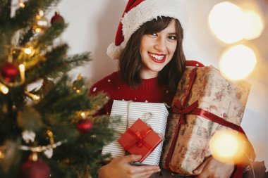 santa şapka şenlikli odasında hediye kutuları altın güzel Noel ağacı ışıkları, yığını tutan mutlu bir genç kadın. mutlu kış tatil atmosferik anlar