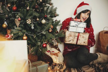 Mutlu Noeller. mutlu kız santa şapka şenlikli odasında birçok hediye kutuları altın güzel Noel ağacı ışıkları, holding. mutlu kış tatil atmosferik anlar. mevsim selam.
