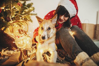 mutlu kız santa şapka şenlikli oda ışıkları ile altın güzel Noel ağacı arka plan üzerinde sevimli köpek ile sarılma içinde. Aile sıcak atmosferik anlar. kış tatili