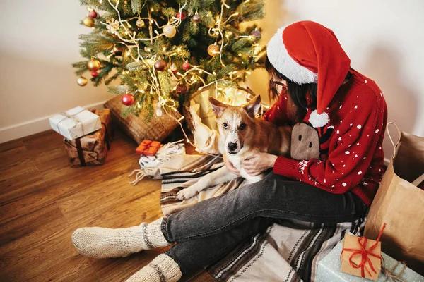 年末年始の家族の暖かい大気瞬間 サンタ帽子を抱いてお祝いルーム灯黄金の美しいクリスマス ツリーの背景にかわいい犬との幸せな女の子 — ストック写真