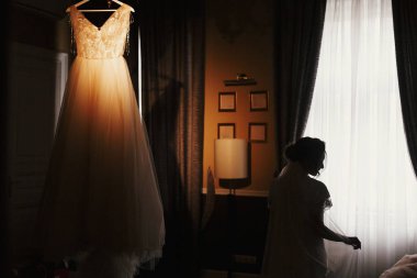Muhteşem gelin siluet duran, şık gelinlik, pencere ışık sabah avize üzerinde asılı. Gelin ve onun güzel gelinlik. Kadın hazırlanıyor