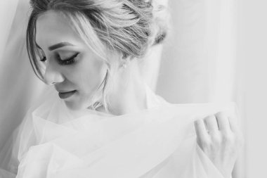 Yumuşak ışık penceresinde yakınındaki sabah poz ipek elbise içinde muhteşem güzel gelin. Holding arka plan beyaz kumaş üzerinde gülümseyen peçe mükemmel makyaj ile mutlu şık gelin