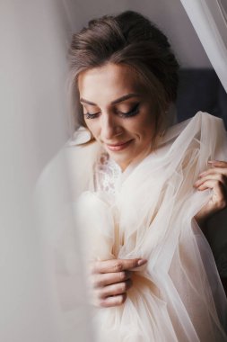 Yumuşak ışık penceresinde yakınındaki sabah poz ipek elbise içinde muhteşem güzel gelin. Mutlu şık gelin peçe tutarak ve gülümseyen mükemmel makyaj ile. Metin için yer.
