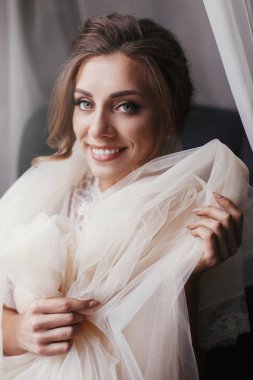 Yumuşak ışık penceresinde yakınındaki sabah poz ipek elbise içinde muhteşem güzel gelin. Mükemmel bir gülümseme ile mutlu seksi gelin. Kadın gülümsüyor ve hazırlanıyor. Metin için yer.