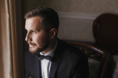 Şık damat siyah takım elbise ve papyon, pencere ışıkta sandalyede oturuyor. Kendinden emin ve mutlu adam portresi. Damat sabah hazırlanıyor