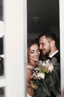Muhteşem gelin ve şık damat yavaşça pencereye sarılma. Kucaklayan ve gülümseyen mutlu evlilik çift. Yeni evliler romantik anlar. Yaratıcı düğün fotoğraf. Kopya alanı