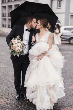 Muhteşem gelin ve yağmurlu sokakta şemsiyesi altında yürüyüş ve öpüşme şık damat. Şehvetli düğün çifti kucaklayan. Yeni evliler romantik anlar. Modern düğün fotoğraf