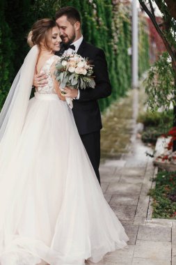 Muhteşem gelin ve şık damat hafifçe yeşil ağaçlar arka plan üzerinde sarılma. Şehvetli düğün çifti kucaklayan. Yeni evliler romantik anlar. Modern düğün fotoğraf