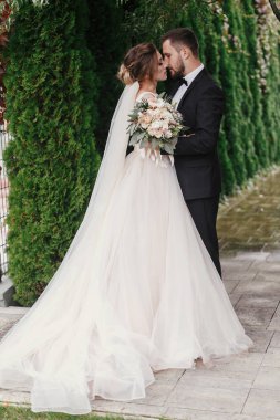 Muhteşem gelin ve şık damat hafifçe yeşil ağaçlar arka plan üzerinde sarılma. Şehvetli düğün çifti kucaklayan. Yeni evliler romantik anlar. Modern düğün fotoğraf