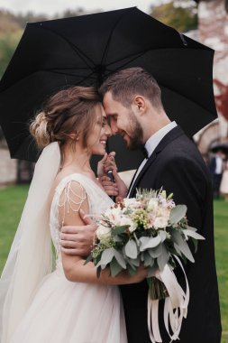Muhteşem gelin ve şık damat yavaşça yağmurlu havada şemsiyesi altında sarılma. Şehvetli düğün çifti kucaklayan. Yeni evliler romantik anlar. Modern düğün fotoğraf