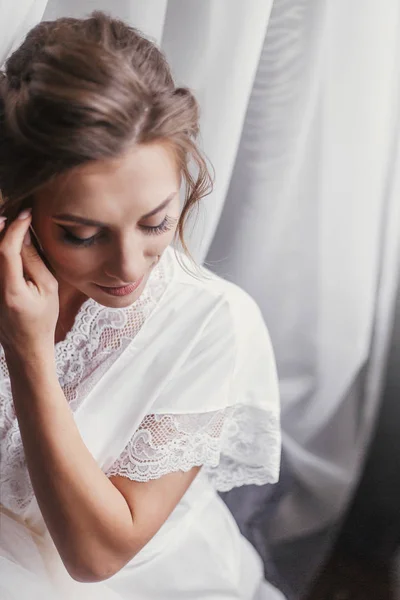 朝の柔らかな光で窓の近くポーズ シルクのローブで豪華な美しい花嫁 白い布の背景に笑みを浮かべてベールで完璧なメイクで幸せなスタイリッシュな花嫁 テキストのためのスペース — ストック写真