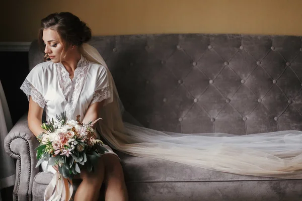 シルクのローブとモダン スタイルのブーケを押しながらスタイリッシュなグレーのソファーに座っていたベールで豪華な美しい花嫁 朝の柔らかな光で窓の近くポーズ完璧なメイクで幸せなスタイリッシュな花嫁 — ストック写真