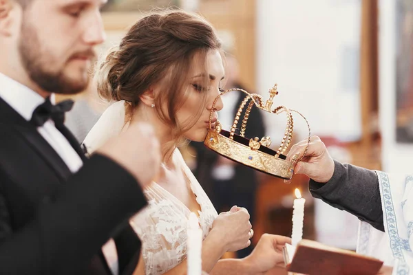 花嫁と花婿の結婚式中に司祭の手からの黄金の冠をキスします 精神的なカップル 結婚式の教会で結婚 感情的なロマンチックな瞬間 — ストック写真