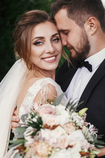 Muhteşem gelin ve yavaşça sarılma ve yeşil ağaçlar arka plan üzerinde gülümseyen şık damat. Şehvetli düğün çifti kucaklayan. Yeni evliler romantik anlar. Düğün fotoğraf