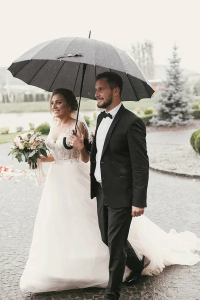Prachtige Bruid Stijlvolle Bruidegom Onder Paraplu Regenachtige Straat Lopen Glimlachen — Stockfoto