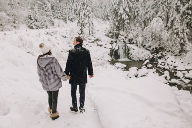 Elele ve kış karlı dağlarda yürüyüş şık çift. Mutlu romantik erkek ve kadın lüks elbiselere rahatlatıcı şelale kar ormanda. Birlikte tatil kaçamağı