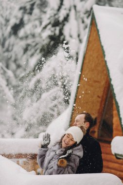 Şık çift ile oynarken kış karlı dağlar arka plan üzerinde ahşap kabin kar. Eğleniyoruz ve köyünde karda gülümseyerek mutlu bir aile. Birlikte tatil kaçamağı