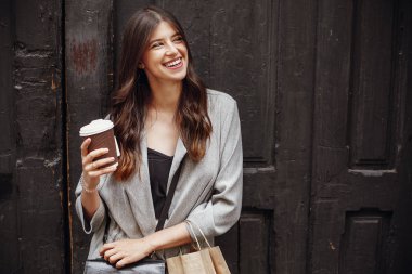 Güzel saç ve kahve Kupası ve kağıt çanta, şehir sokak eski ahşap kapıda ayakta tutan bir gülümseme ile şık hipster kız. Vaktinizi ve alışveriş muhteşem mutlu genç kadın