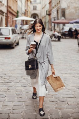 Gülümseyen ve şehir sokak yürüyüş fincan kahve ve çanta, tutan güzel genç kadın. Güzel saç ve zaman zevk ve alışveriş mükemmel bir gülümseme ile şık mutlu hippi kız